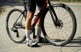 Un ciclista con calcetines blancos y zapatillas de ciclismo negras posa junto a una bicicleta de carretera BMC negra y roja, equipada con ruedas Zipp y neumáticos Schwalbe Pro One Aero, en una carretera asfaltada.