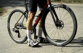 Un ciclista posa junto a una bicicleta de carretera con ruedas Zipp en una carretera asfaltada; la rueda delantera lleva un neumático Schwalbe Pro One Aero, y él lleva pantalones cortos negros, calcetines blancos y zapatillas de ciclismo.