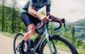 Un hombre con casco y atuendo ciclista monta en una bicicleta de carretera SCOTT Addict en un sendero al aire libre con árboles y colinas de fondo.