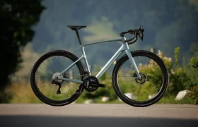 Una bicicleta de carretera SCOTT Addict con manillar rebajado, frenos de disco y neumáticos finos está aparcada en una superficie pavimentada con vegetación y colinas al fondo.