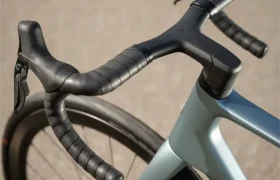 Primer plano del manillar y la rueda delantera de una moderna bicicleta de carretera SCOTT Addict, mostrando los puños del manillar envueltos y las manetas de freno integradas sobre una superficie pavimentada.