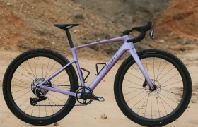 Una bicicleta gravel SCOTT Addict Gravel 2027 de color morado, con neumáticos negros, está aparcada sobre terreno arenoso, con un paisaje rocoso y montañoso al fondo.