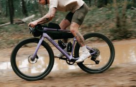 Una persona que circula en una bicicleta SCOTT Addict Gravel 2027 de color morado por un sendero polivalente y embarrado en un bosque; lleva casco, camiseta beige, pantalones cortos verdes y zapatillas blancas. Se le ven tatuajes en los brazos y las piernas.