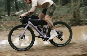 Una persona que va en una bicicleta SCOTT Addict Gravel 2027 de color morado por un sendero embarrado que atraviesa un bosque, vestida con pantalones cortos, una camiseta ligera y gafas de sol.