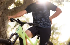 Una persona que lleva casco y gafas de sol posa al aire libre junto a una bicicleta SCOTT Strike 2027 de color verde, con la luz del sol brillando a sus espaldas a través de los árboles. El potente motor Bosch CX de la bicicleta garantiza la comodidad en cada recorrido.