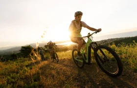 Tres ciclistas suben en sus bicicletas de montaña SCOTT Strike 2027 por un sendero de tierra al atardecer, disfrutando tanto de la comodidad como de la potente asistencia que proporciona el motor Bosch CX, con un terreno cubierto de hierba y un paisaje pintoresco al fondo.