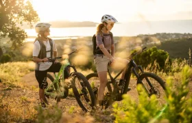 Dos personas con casco posan junto a sus bicicletas de montaña SCOTT Strike 2027 en un sendero de tierra, con vistas a un paisaje en el que se aprecian vegetación y agua al fondo durante la puesta de sol. Gracias al potente motor Bosch CX y a su excepcional comodidad, están listas para afrontar cualquier aventura.