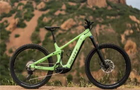 Una bicicleta de montaña SCOTT Strike 2027 de color verde, con una suspensión de 150 mm y un potente motor Bosch CX, está aparcada en una carretera asfaltada, con colinas rocosas y un cielo nublado al fondo.