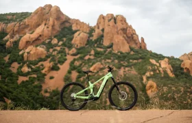 Una bicicleta de montaña SCOTT Strike 2027 de color verde, con una suspensión de 150 mm, se encuentra en un camino asfaltado con grandes colinas rocosas y vegetación al fondo, bajo un cielo nublado.
