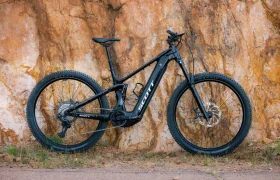 Una bicicleta de montaña eléctrica SCOTT Strike 2027 de color negro, con neumáticos anchos y un potente motor Bosch CX, está aparcada en posición vertical junto a una pared rocosa de color naranja y de aspecto rugoso, en un camino de tierra.