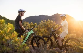 Dos personas con casco están sentadas en sus bicicletas de montaña SCOTT Strike 2027, disfrutando de la comodidad que les proporciona el avanzado motor Bosch CX mientras se encuentran cara a cara en un sendero, con vegetación y colinas al fondo durante la puesta de sol.