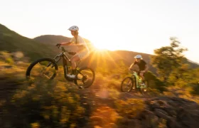 Dos ciclistas, con casco, recorren en sus bicicletas de montaña SCOTT Strike 2027 —equipadas con un potente motor Bosch CX y una suspensión de 150 mm— un sendero rocoso al atardecer, con colinas y vegetación al fondo.