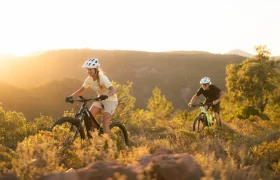 Dos personas con casco circulan en bicicletas de montaña SCOTT Strike 2027, equipadas con un potente motor Bosch CX, por un sendero que atraviesa colinas bañadas por el sol y la vegetación, disfrutando de una comodidad excepcional con las montañas como telón de fondo.