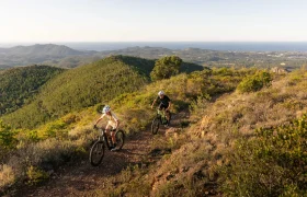 Dos personas recorren en sus bicicletas de montaña SCOTT Strike 2027 un sendero de tierra que atraviesa un terreno verde y accidentado, con vistas lejanas al mar y un cielo despejado, disfrutando tanto de la comodidad como de la potencia que ofrece el motor Bosch CX.