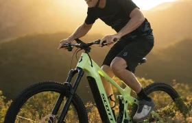 Un hombre con casco pedalea en una bicicleta de montaña SCOTT Strike 2027, equipada con un potente motor Bosch CX, por un sendero con colinas y la luz del sol al fondo, disfrutando tanto del rendimiento como de la comodidad durante su recorrido.