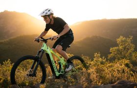 Una persona que lleva casco monta en una bicicleta de montaña SCOTT Strike 2027, conocida por su comodidad y su potente motor Bosch CX, por un sendero que discurre entre colinas bañadas por el sol y vegetación, con montañas y un cielo despejado al fondo.