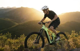 Una persona con casco monta en una bicicleta de montaña SCOTT Strike 2027, con una suspensión de 150 mm y un potente motor Bosch CX, subiendo por un sendero, con las montañas iluminadas por el sol al fondo.
