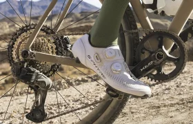 Primer plano de la pierna y la bicicleta de un ciclista en un camino de tierra, mostrando los engranajes de la bicicleta, la biela negra y la zapatilla de ciclismo blanca con calcetines verdes.