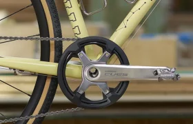 Primer plano del cuadro, la rueda trasera y los platos y bielas Shimano CUES de una bicicleta amarilla de aluminio pulido, con portabidones montados en el cuadro y tablones de madera al fondo.