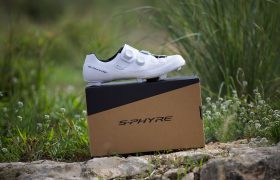 Una Zapatillas Shimano RX910 S-Phyre blanca para ciclismo de gravilla con dos esferas se muestra encima de una caja S-Phyre marrón al aire libre, con hierba y rocas de fondo.