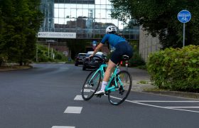 Un ciclista con traje y casco azules monta en una bicicleta de carretera verde azulado en una calle de la ciudad cerca de una señal de "sólo bicicletas" y un edificio rotulado "FASHION CENTER".