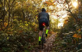 Un ciclista con calcetines amarillo neón monta en bicicleta por un estrecho sendero boscoso, con la luz del sol filtrándose entre los árboles y un SIGMA NYTE CLIP bien sujeto a su equipo para mayor visibilidad.