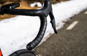 Primer plano del manillar negro de una bicicleta de carretera, envuelto con cinta Silca Nastro Cuscino, y de la palanca de freno, en una carretera asfaltada con nieve en los márgenes, lo que pone de relieve la excepcional comodidad y el tacto superior en cada salida.