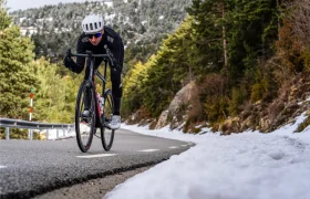 Un ciclista, con casco y gafas de sol, sube por una carretera de montaña, disfrutando de una comodidad excepcional y un tacto mejorado gracias a la cinta de manillar Silca Nastro Cuscino, con nieve a los lados de la carretera y árboles al fondo.