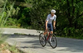 Un ciclista, con casco blanco y equipación de ciclismo, pedalea en una bicicleta de carretera roja y negra, disfrutando de una mayor comodidad y un tacto superior gracias a la cinta de manillar Silca Nastro Cuscino, mientras sube por una carretera asfaltada rodeada de vegetación.