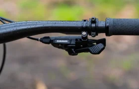 Primer plano de un manillar de bicicleta negro con empuñadura texturizada y cambio SRAM Eagle, con gotas de agua brillando sobre un fondo natural difuminado. Perfecto para quienes aprecian la precisión y la fiabilidad de los cambios mecánicos.
