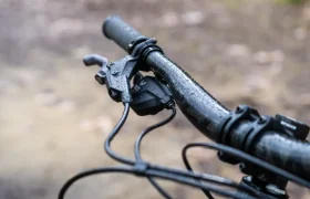 Primer plano de un manillar de bicicleta mojado, equipado con palancas de freno SRAM Eagle 90 y cables visibles sobre un fondo de camino exterior borroso.