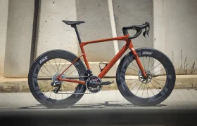 Una moderna bicicleta de carretera con cuadro rojo y ruedas de sección profunda, diseñada para el máximo rendimiento, se alza orgullosa contra una pared de hormigón.