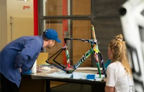 Dos personas examinan un colorido cuadro de bicicleta BMC VAR0 en una mesa de trabajo dentro de un taller o estudio.