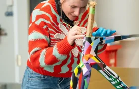 Una mujer que viste un suéter rojo estampado pinta un diseño geométrico colorido en una pequeña escultura de un animal en un espacio de trabajo interior en BMC VAR0.