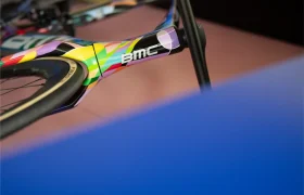 Primer plano de un cuadro de bicicleta BMC VAR0 con un diseño geométrico colorido, centrándose en la horquilla delantera y el neumático, parcialmente oscurecidos por un objeto azul.