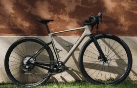 Una moderna bicicleta de carretera Van Rysel EDR-G con manillar de carretera está aparcada sobre la hierba verde, frente a una pared con textura en tonos marrones y blancos.