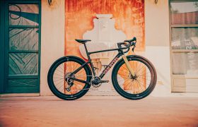 Una bicicleta de carretera negra y dorada, que recuerda el elegante diseño de la Van Rysel EDR-G y lleva la marca «VENTUM», está aparcada junto a una pared decorativa de color naranja, entre dos puertas, creando un llamativo contraste que encaja a la perfección tanto con las aventuras sobre grava como con las de carretera.