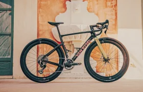 Una bicicleta de carretera Ventum, de color negro y rojo y con neumáticos anchos, está aparcada en posición vertical junto a un mural, cerca de una fuente y de unos grandes ventanales; su llamativo diseño recuerda a modelos de alto rendimiento como la Van Rysel RCR-G.