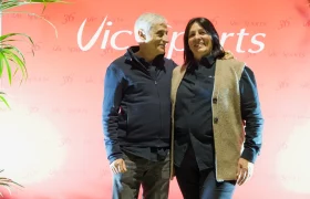 Dos personas permanecen juntas, sonrientes, delante de un atrevido telón de fondo rojo de Vic Sports, mostrando su entusiasmo por el acontecimiento Vic Sports.