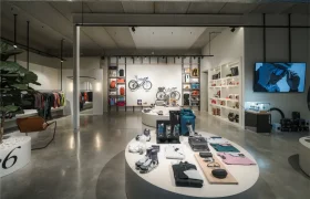 Interior de tienda moderna con mesas redondas de exposición de ropa y accesorios de Vic Sports, estanterías con bolsos y una bicicleta en la pared del fondo, y una pantalla que muestra un anuncio.