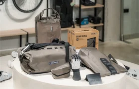 Un expositor de bolsas y accesorios Ortlieb de color caqui, que incluye una bolsa de viaje, un bolso tote, guantes y una bolsa para bicicletas en caja, dispuestos sobre una mesa redonda blanca en la tienda de Vic Sports.
