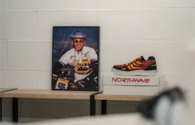 Una foto enmarcada de un ciclista y unas zapatillas Northwave rojas y amarillas descansan sobre dos cajas apiladas de Northwave de Vic Sports, expuestas en una estantería de madera contra una pared blanca.