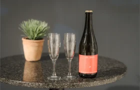 Una botella de bebida espumosa con etiqueta roja y dos copas de champán vacías sobre una mesa redonda oscura, con una gorra de Vic Sports descansando cerca y una maceta al fondo.