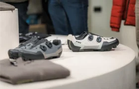 Dos zapatillas de ciclismo Vic Sports con cierre de esfera se exponen sobre una superficie blanca, con prendas de vestir dobladas visibles en las proximidades.