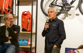 Dos hombres hablan en un evento deportivo cubierto de Vic Sports cerca de estanterías con bolsas y una bicicleta montadas en la pared; uno de ellos sujeta un micrófono mientras el otro se sienta y sonríe.