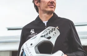 Un hombre que lleva una chaqueta negra de Peugeot sostiene al aire libre un casco blanco de Peugeot Cycles, con bicicletas visibles al fondo, lo que pone de manifiesto el estilo emblemático de esta marca histórica.