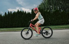 Una niña con casco conduce una bicicleta rosa WOOM 4 Explore por una carretera pavimentada, con árboles y un cielo nublado de fondo.