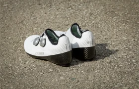 Un par de Zapatillas Q36.5 Unique Pro blancas con suelas negras y cierres de dial descansa sobre un asfalto rugoso, listas para ofrecer el máximo rendimiento.