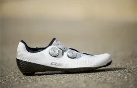 Una zapatilla de ciclismo Zapatillas Q36.5 Unique Pro blanca con dos cierres de esfera y una suela de carbono negra descansa sobre una superficie texturizada, mostrando a la vez un diseño moderno y rendimiento. El lateral muestra el logotipo "Q36.5.