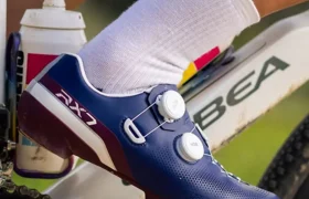 Primer plano del pie de un ciclista que lleva unas zapatillas azules Shimano RX710 en el pedal de una bicicleta, en las que se aprecia la tecnología de competición; también se ven un bidón y parte del cuadro blanco de una bicicleta ORBEA, ideal para cualquier aventura de gravel.